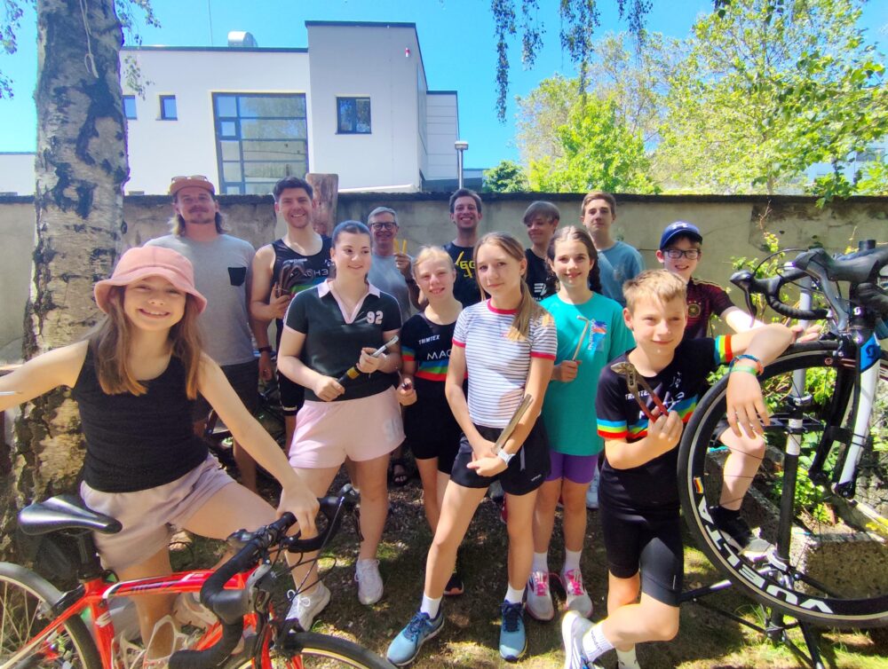 Gruppenfoto der Triathlon-Jugend von MACH3 Köln e.V. beim DIY-Rennrad-Workshop vor der Werkstatt von Velonia. Kinder, Jugendliche und Trainer lächeln mit Werkzeug und Fahrrädern fröhlich in die Kamera.