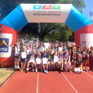 Über 30 Triathlon Kinder Köln stehen stolz mit Urkunden und Hawaii-Ketten unter dem großen Zielbogen des NRWTV.