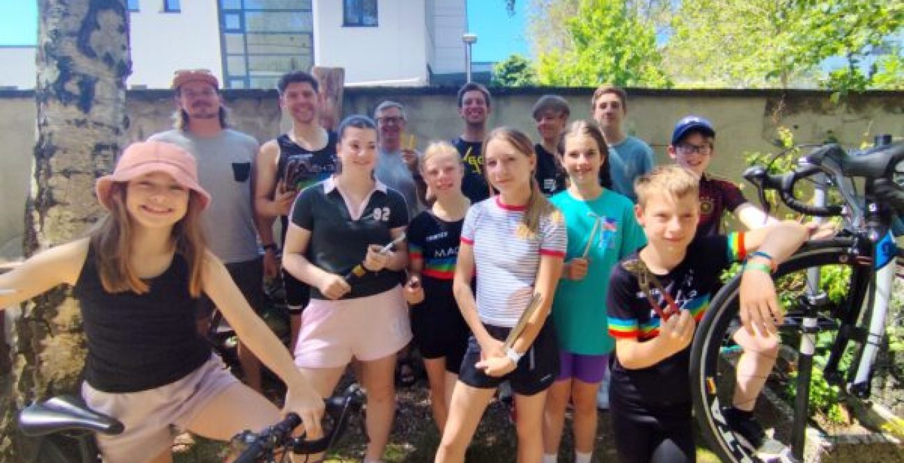 Gruppenfoto der Triathlon-Jugend von MACH3 Köln e.V. beim DIY-Rennrad-Workshop vor der Werkstatt von Velonia. Kinder, Jugendliche und Trainer lächeln mit Werkzeug und Fahrrädern fröhlich in die Kamera.