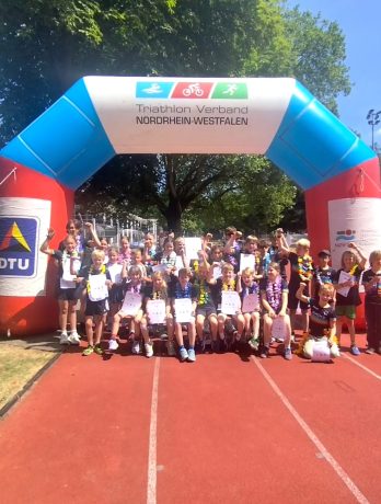 Über 30 Triathlon Kinder Köln stehen stolz mit Urkunden und Hawaii-Ketten unter dem großen Zielbogen des NRWTV.