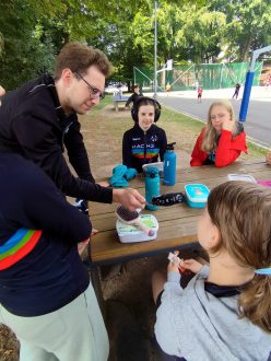 Ein WDR-Journalist interviewt mit einem Mikrofon aufmerksame Kinder am Holztisch während des MACH3 Feriencamps.